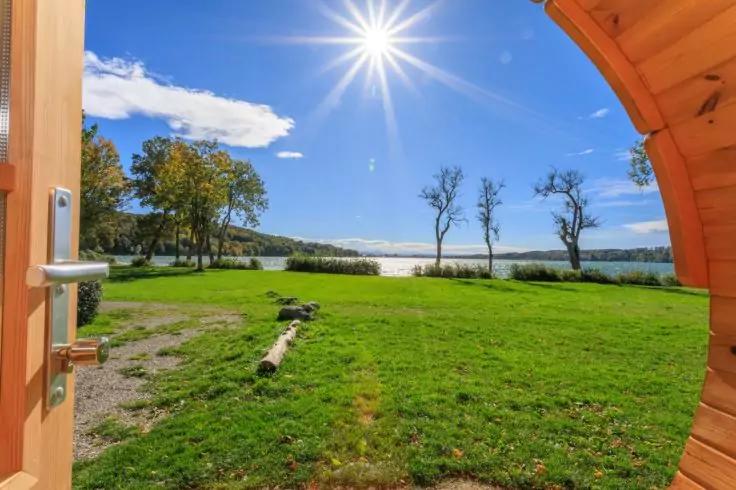 Runde Holz&ouml;ffnung mit T&uuml;rgriff links und gebogener Holzwand rechts, gr&uuml;ne Wiese mit liegendem Baumstamm, See am Horizont, B&auml;ume am Ufer, strahlende Sonne am blauen Himmel mit wenigen Wolken
