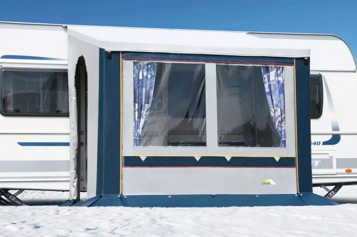 Wei&szlig;er Wohnwagen mit blau-wei&szlig;em Anbau mit gro&szlig;em Fenster und gemusterten Gardinen; verschneiter Boden und blauer Himmel