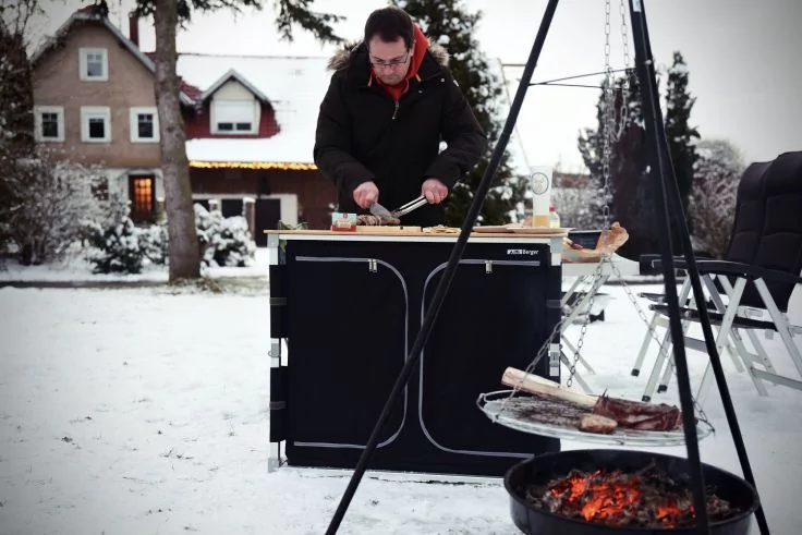 Person in Winterjacke steht hinter einer schwarzen Kochbox im Schnee und schneidet auf einem Holzbrett; im Vordergrund eine Feuergrube mit gl&uuml;henden Kohlen und Grillrost mit Fleischst&uuml;cken; rechts Klappst&uuml;hle; H&auml;user und B&auml;ume im Hintergrund