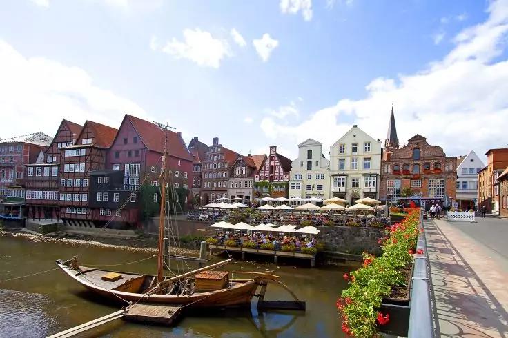 Mehrst&ouml;ckige Fachwerk- und Ziegelh&auml;user am Ufer eines Kanals, mehrere h&ouml;lzerne Boote am Wasser, Au&szlig;enterrassen mit Sonnenschirmen und sitzenden Personen, Fu&szlig;g&auml;ngerbr&uuml;cke mit Blumenk&auml;sten, teilweise bew&ouml;lkter Himmel