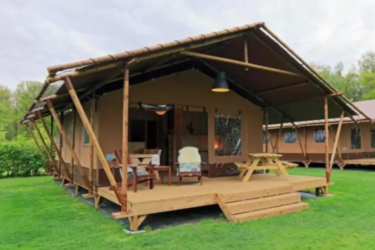 Holzh&uuml;tte auf erh&ouml;hter Plattform mit &uuml;berdachter Veranda, zwei Sesseln, Holztisch mit B&auml;nken, h&auml;ngende Lampe, Stufen, Rasen und B&auml;ume im Hintergrund