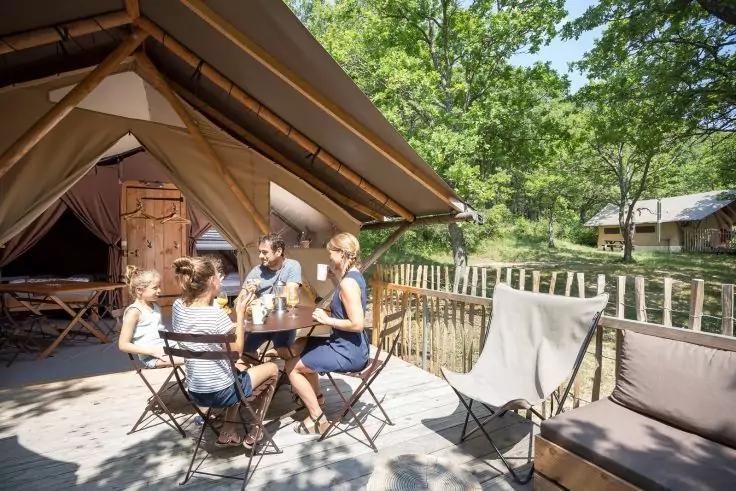 Vier Personen sitzen an einem Tisch auf einer Holzterrasse vor einem Zelt; B&auml;ume und ein weiteres Zelt im Hintergrund