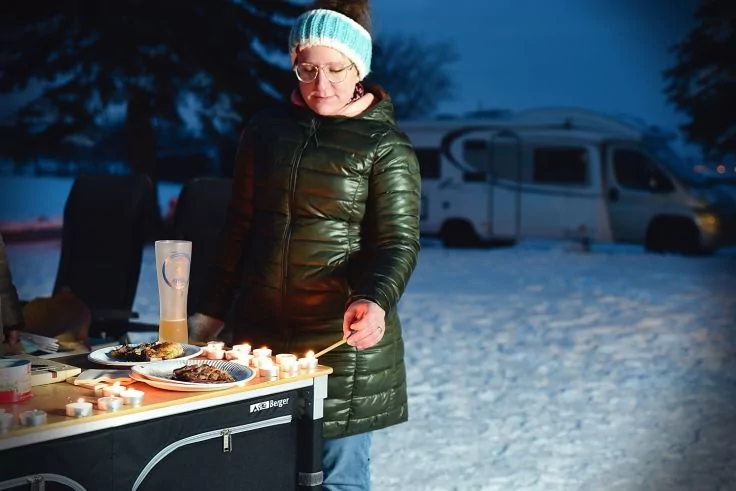 Person in gr&uuml;nem Steppmantel und hellblauem Stirnband steht nachts im Schnee an einem Tisch mit brennenden Teelichtern, Tellern mit Essen und einer Thermoskanne; Wohnmobil im Hintergrund