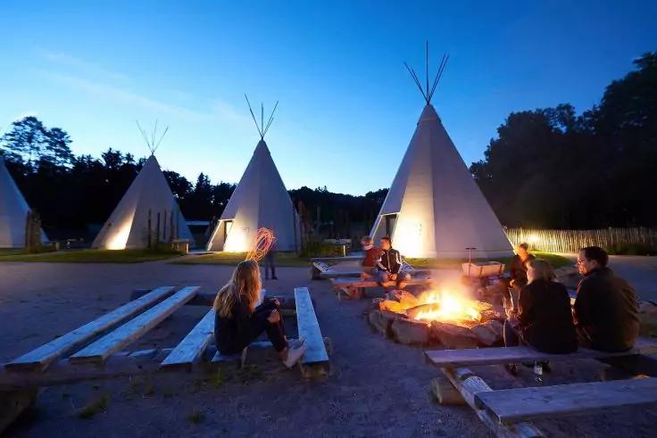 Drei beleuchtete Tipis bei D&auml;mmerung, Personen sitzen auf Holzb&auml;nken um ein Lagerfeuer, B&auml;ume am Horizont