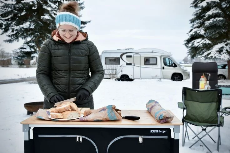 Person in Winterjacke und Stirnband schneidet Brot auf Campingtisch; Wohnmobil und Klappstuhl im Schnee