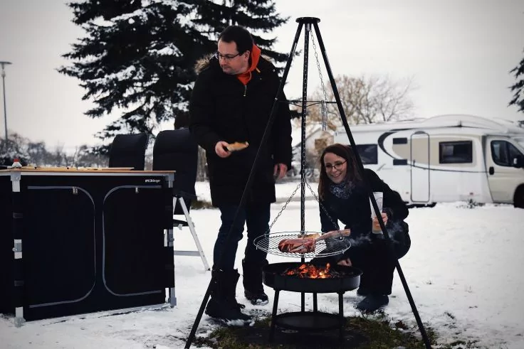 Zwei Personen bereiten auf einem Dreibeingrill &uuml;ber offenem Feuer im Schnee W&uuml;rstchen zu; Wohnwagen und Baum im Hintergrund
