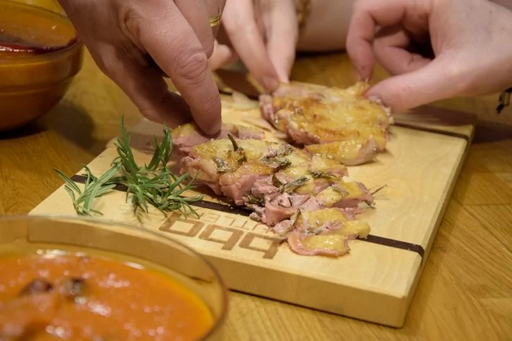 H&auml;nde nehmen d&uuml;nne Fleischscheiben von einem Holzbrett mit einem Zweig Rosmarin, daneben Sch&uuml;sseln mit roter Sauce