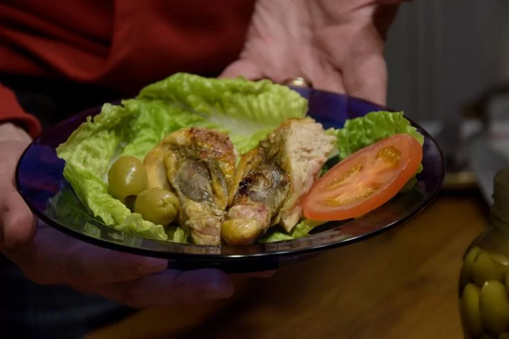 Blaue Glasschale, von zwei H&auml;nden gehalten, mit einem Salatblatt, aufgeschnittenem gebr&auml;untem Gefl&uuml;gelst&uuml;ck, gr&uuml;nen Oliven, einer Tomatenscheibe und Brot; Tisch und ein Glas mit Oliven im Hintergrund