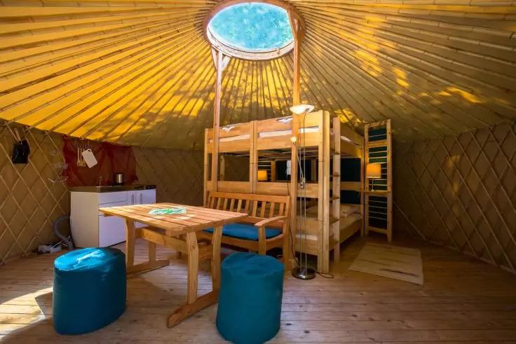 Runder Innenraum mit Holzboden, rundem Oberlicht, h&ouml;lzernem Etagenbett, Holztisch mit zwei blauen Hockern, kleinem wei&szlig;em K&uuml;hlschrank und Regalen