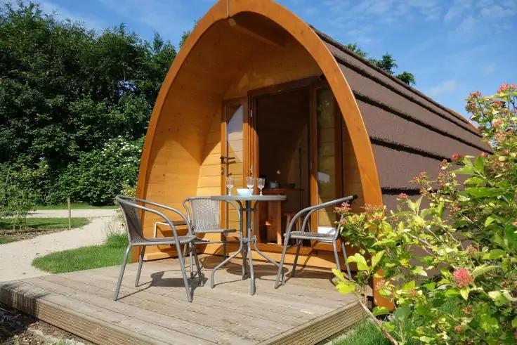 Geschwungene Holz-Pod mit offener T&uuml;r, Holzdeck mit rundem Tisch und zwei Metallst&uuml;hlen, Str&auml;ucher und B&auml;ume im Hintergrund