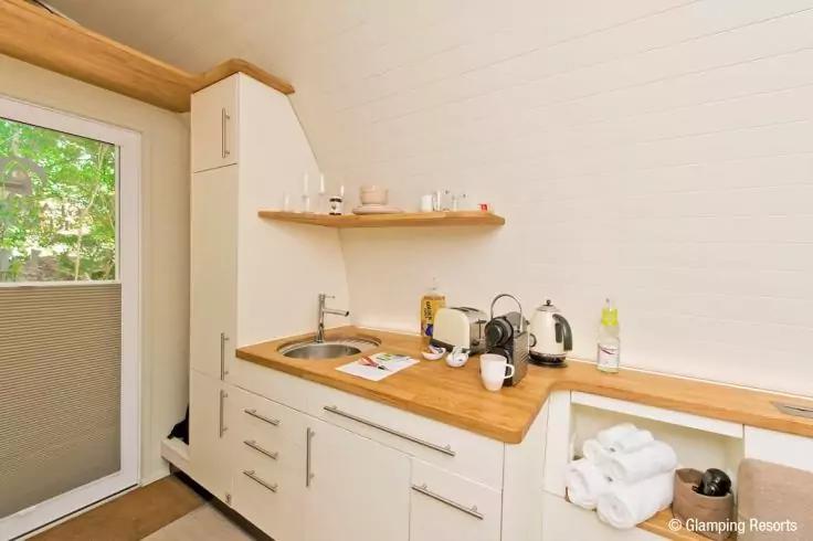 K&uuml;chenzeile mit Holzarbeitsplatte, Sp&uuml;le, Wasserkocher, Tassen, Regal mit Gl&auml;sern, aufgerollten Handt&uuml;chern und einer T&uuml;r mit Rollo