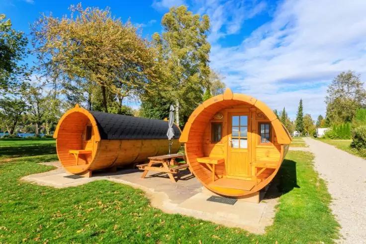Zwei runde Holzh&auml;user in Fassform auf Betonplatten nebeneinander, mit einem Picknicktisch dazwischen, umgeben von Gras, B&auml;umen und einem Weg, blauer Himmel mit Wolken