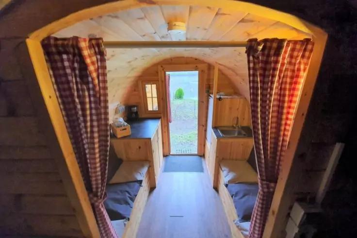 Enger Innenraum aus Holz mit zwei Einzelbetten an den Seiten, karierten Vorh&auml;ngen, K&uuml;chenzeile mit Sp&uuml;le und Wasserkocher und ge&ouml;ffneter T&uuml;r nach drau&szlig;en