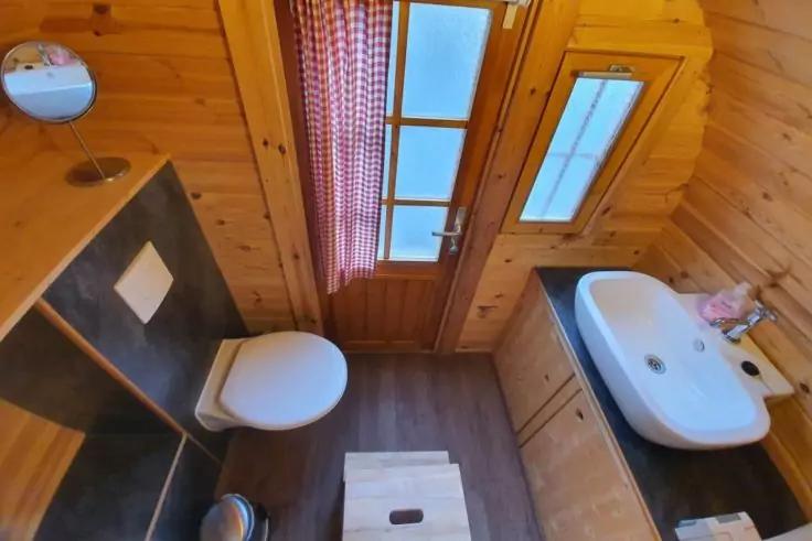 Kleines holzverkleidetes Badezimmer mit Toilette links, Waschbecken rechts mit Seifenflasche, Holzt&uuml;r mit mattierten Glasscheiben und kariertem Vorhang, runder Spiegel an der Wand, kleiner M&uuml;lleimer und Holzhocker auf dem Boden