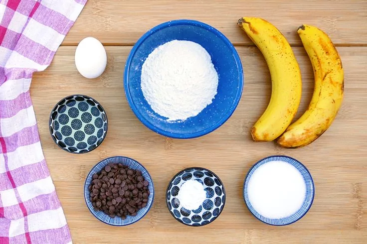 Zwei Bananen, ein Ei, blaue Sch&uuml;sseln mit Mehl, Zucker und Schokost&uuml;ckchen, zwei kleine gemusterte Schalen und ein rosa-wei&szlig; kariertes Tuch auf Holzuntergrund