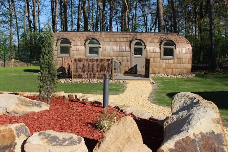 Gebogenes Holzgeb&auml;ude mit drei halbrunden Fenstern und einer T&uuml;r; Kiesweg von Vordergrund zum Eingang; Vorgarten mit gro&szlig;en Felsen, rotem Mulch und B&auml;umen im Hintergrund