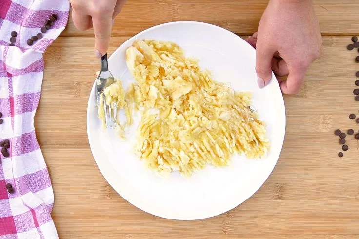 Zwei H&auml;nde zerdr&uuml;cken mit einer Gabel eine gesch&auml;lte Banane auf einem wei&szlig;en Teller; Teller steht auf Holzuntergrund, links ein lila kariertes Tuch, rechts verstreute Kaffeebohnen