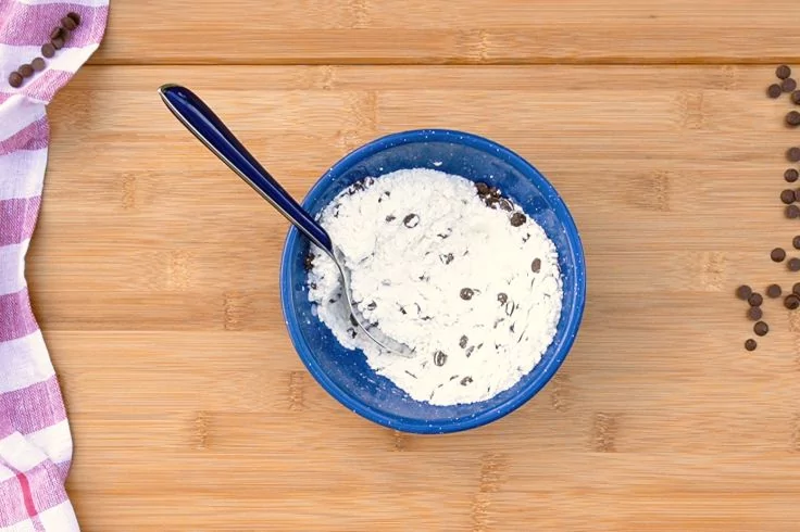 Blaue Schale mit wei&szlig;er Masse und Schokost&uuml;ckchen, L&ouml;ffel darin, auf Holzbrett; links gestreiftes Tuch, rechts verstreute Schokost&uuml;ckchen