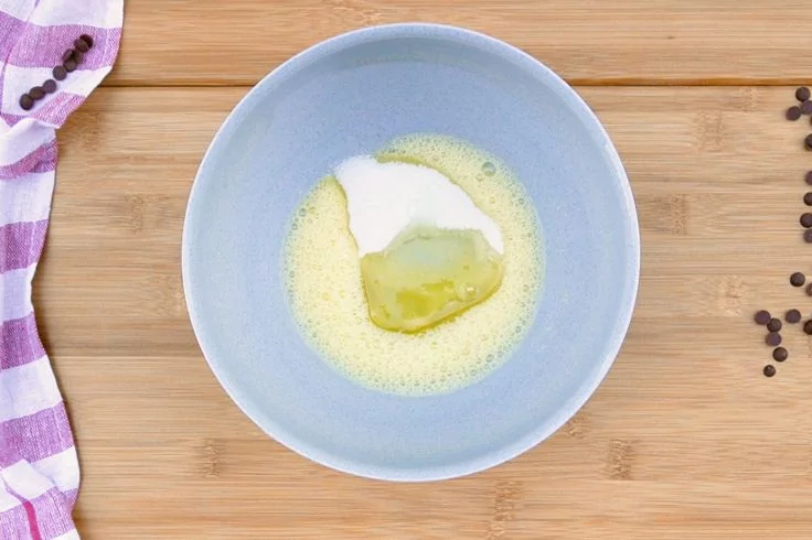 Hellblaue Sch&uuml;ssel mittig auf Holzbrett, darin gelblich sch&auml;umende Fl&uuml;ssigkeit und ein wei&szlig;er Klumpen; links lila-wei&szlig; gestreiftes Tuch, rechts verstreute kleine dunkle K&uuml;gelchen