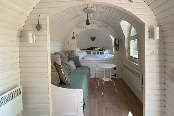 Schmaler, wei&szlig; gestrichener Innenraum mit gew&ouml;lbter Decke; links gepolsterte Sitzbank mit Kissen, mittig kleiner runder wei&szlig;er Tisch, rechts Fenster mit Rolll&auml;den und Heizk&ouml;rper, hinten Liege mit herzf&ouml;rmiger Wanddekoration, h&auml;ngende Lampen, Holzbode