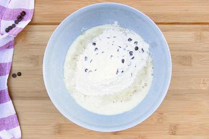 Hellblaue Sch&uuml;ssel mit wei&szlig;em Pulver, heller Fl&uuml;ssigkeit und kleinen dunklen St&uuml;ckchen auf Holzuntergrund; lila-wei&szlig; gestreiftes K&uuml;chentuch links