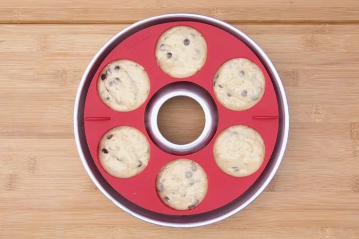 Roter runder Silikon-Backring mit sieben runden Mulden, jeweils gef&uuml;llt mit hellem Teig mit Schokost&uuml;ckchen, auf Holzuntergrund