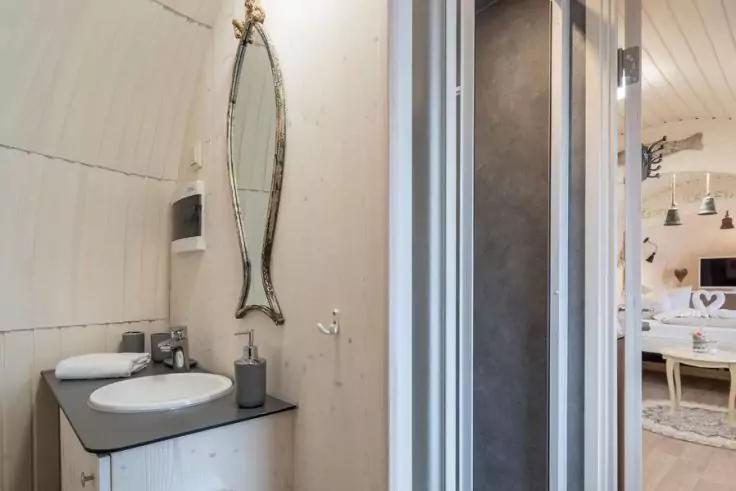 Waschbecken auf dunkler Arbeitsfl&auml;che, Seifenspender, ovaler Wandspiegel mit Rahmen, helle Holzpaneele, teilweise ge&ouml;ffnete Faltt&uuml;r, angrenzendes Zimmer mit Bett und H&auml;ngeleuchten