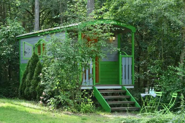 Gr&uuml;n gestrichener Holzwohnwagen mit Treppe und kleinem Verandavorbau, umgeben von B&auml;umen und Str&auml;uchern, davor gr&uuml;ner Klapptisch und St&uuml;hle auf Gras