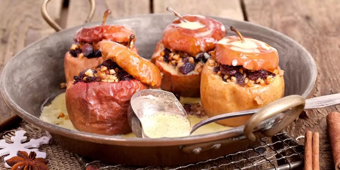 Gef&uuml;llte &Auml;pfel mit N&uuml;ssen und getrockneten Beeren in einer Pfanne mit So&szlig;e und einem L&ouml;ffel, Zimtstangen daneben