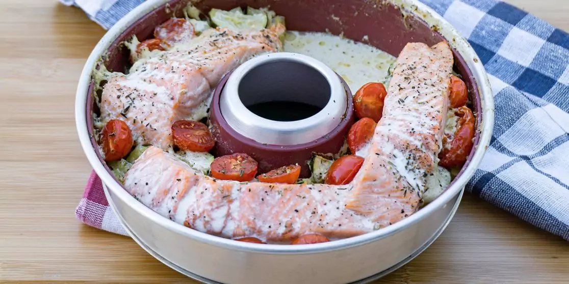 Rundes Rohrbackformblech mit zentralem Rohr, rohe Lachsfilets in einem Ring, halbierte Kirschtomaten und gr&uuml;ne Kr&auml;uterflocken auf einer wei&szlig;-cremefarbenen Masse; Blech auf Holzuntergrund, karierte K&uuml;chenhandt&uuml;cher daneben