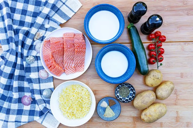 Holzuntergrund mit blau-weiß kariertem Küchentuch, Teller mit Lachsfilets, zwei blaue Schalen mit weißer Flüssigkeit, Schale mit geriebenem Käse, zwei kleine Schalen mit Gewürzen, Zucchini, drei Kartoffeln, Rispe Kirschtomaten, Salz- und Pfefferstreu