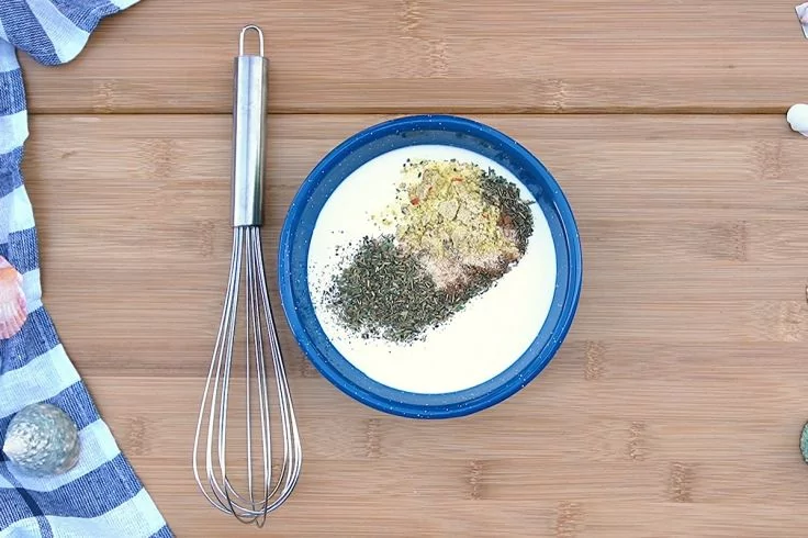 Blaue Schüssel mit weißer Flüssigkeit und drei Gewürzstreifen darauf; Metall-Schneebesen links; kariertes Tuch links auf Holzbrett