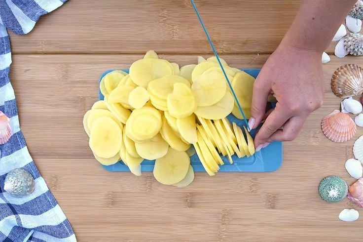 Hand schneidet dünne gelbe Scheiben auf einem blauen Schneidebrett auf einer Holzoberfläche; Muscheln rechts, gestreiftes Tuch links