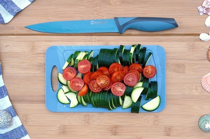 Blaues Schneidebrett mit in Scheiben geschnittener Zucchini und halbierten Kirschtomaten, darüber ein Küchenmesser auf Holzuntergrund, Stofftuch und Muscheln am Rand