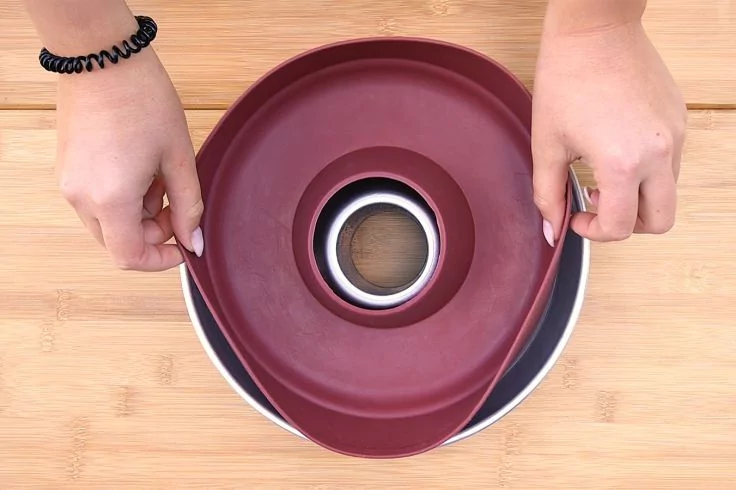 Zwei Hände halten eine flexible dunkelrote Silikonform über einer runden Metallbackform mit mittiger Öffnung auf einem Holzbrett