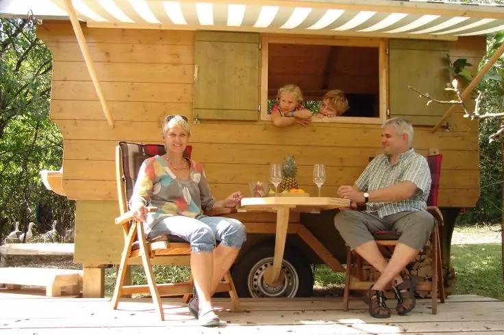 Zwei Erwachsene sitzen an einem kleinen Tisch vor einem h&ouml;lzernen Bauwagen; zwei Kinder lehnen aus einem offenen Fenster. Flasche und Gl&auml;ser auf dem Tisch, B&auml;ume im Hintergrund