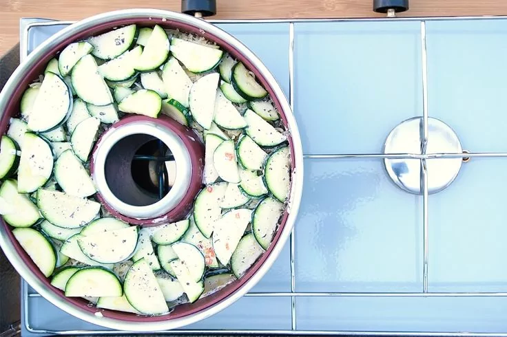 Aufsicht auf eine runde Napf-/Kranzform mit Loch in der Mitte, gefüllt mit dünn geschnittenen Zucchinischeiben in kreisförmiger Anordnung, gewürzt, auf einem Herd mit Metallgitter