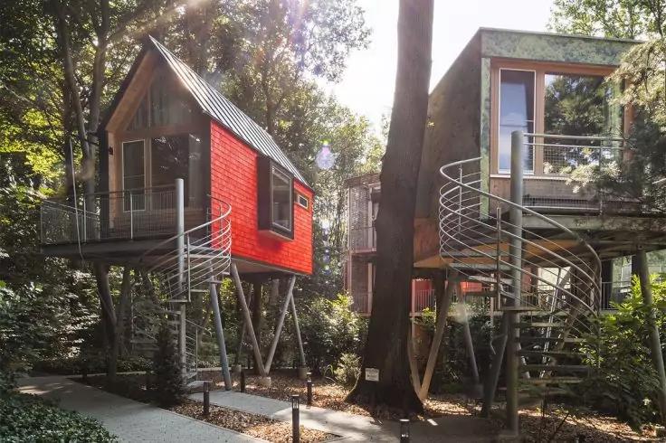 Zwei moderne Baumh&auml;user auf Stelzen zwischen B&auml;umen, links rot, rechts grau mit spiralf&ouml;rmiger Au&szlig;entreppe