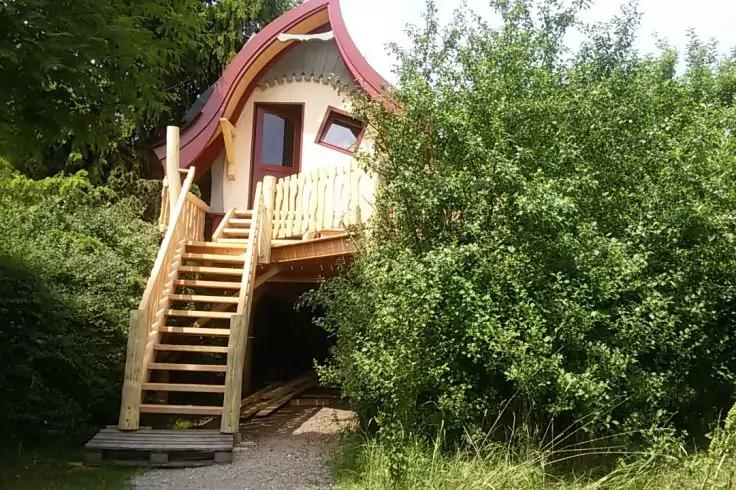 Erh&ouml;hte Holzh&uuml;tte mit rotem, gebogenem Dach, Holztreppe und Gel&auml;nder, umgeben von gr&uuml;nen B&uuml;schen und B&auml;umen, Kiesweg im Vordergrund