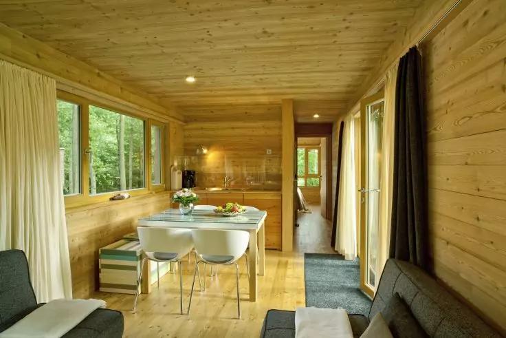 Innenraum einer Holzh&uuml;tte mit Holzverkleidung an W&auml;nden, Decke und Boden, Esstisch mit vier wei&szlig;en St&uuml;hlen, gedeckter Tisch und Blumen, gro&szlig;e Fenster mit B&auml;umen, Sofas, offener Durchgang zum Flur