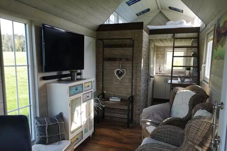 Innenraum eines kompakten Wohnraums mit zwei gepolsterten Sesseln rechts, Kommode mit an der Wand montiertem Fernseher links, Regal mit Herzdekoration in der Mitte, Leiter zu einem Hochbett, Fenster mit Blick auf eine Wiese, Holzboden und schr&auml;ges Ho