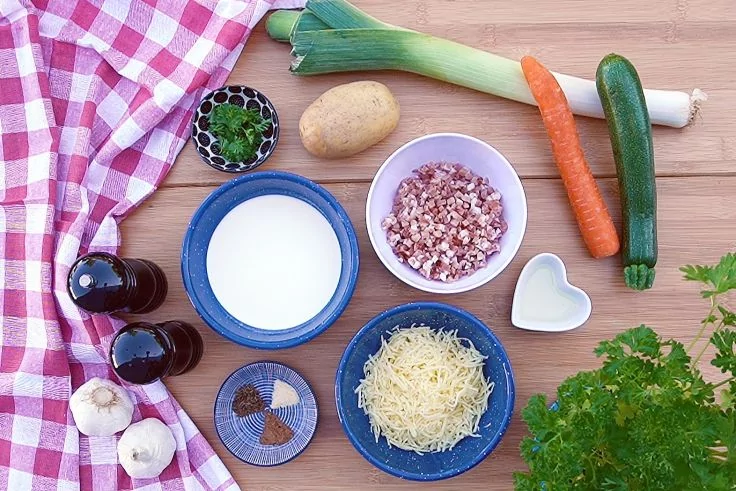 Auf Holzbrett: rosa-wei&szlig; kariertes Tuch, zwei Knoblauchknollen, zwei dunkle Glasflaschen, kleine Schale mit gr&uuml;nen Kugeln, blaue Schale mit wei&szlig;er Fl&uuml;ssigkeit, blaue Schale mit geriebenem gelben K&auml;se, kleine Schale mit gew&uuml;rfelten rosa-wei&szlig;en St&uuml;cken