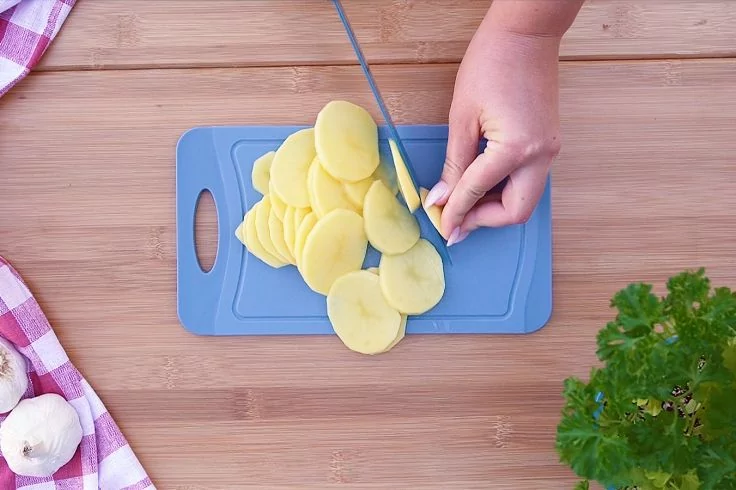 Hand schneidet Kartoffeln in d&uuml;nne Scheiben auf einem blauen Schneidebrett auf einem Holztisch; rechts Petersilie, links Knoblauchknollen und ein kariertes Tuch