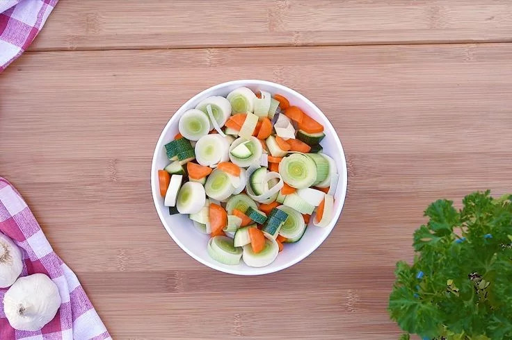 Wei&szlig;e Sch&uuml;ssel mit Lauchringen, Karotten- und Zucchinist&uuml;cken auf Holzoberfl&auml;che; links kariertes Tuch mit Knoblauchknollen, rechts Bund Petersilie