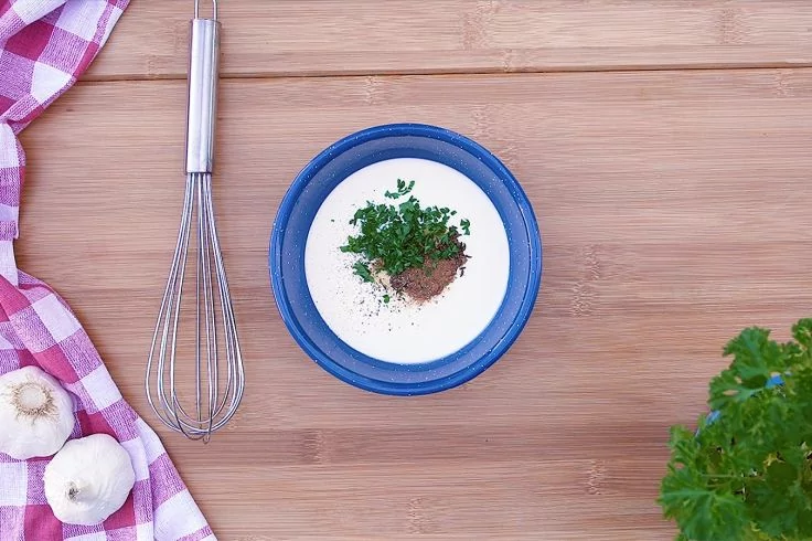 Blaue Schale mit wei&szlig;er cremiger Masse, in der Mitte gehackte Kr&auml;uter und braunes Gew&uuml;rz; links Schneebesen, Knoblauchknollen und rot-wei&szlig; kariertes Tuch; rechts Petersilie auf Holzuntergrund