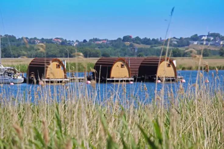 Drei fassf&ouml;rmige schwimmende H&uuml;tten auf einem See, Schilf im Vordergrund, Ufer mit H&auml;usern im Hintergrund, blauer Himmel