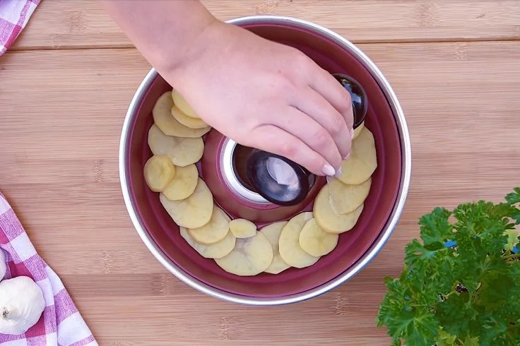 Hand legt Kartoffelscheiben in eine Gugelhupf-Backform auf einem Holzbrett; rechts Topf mit Petersilie, links Knoblauch und rot-wei&szlig;es Tuch