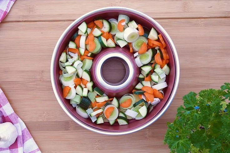 Runde Backform mit Loch, gef&uuml;llt mit geschnittenen Zucchini-, Karotten- und Lauchst&uuml;cken auf Holzbrett; links kariertes K&uuml;chentuch, rechts Petersilie