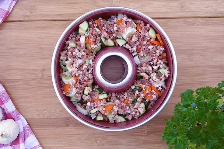 Rundes Metallgef&auml;&szlig; mit Loch in der Mitte, gef&uuml;llt mit feinen K&ouml;rnern und gew&uuml;rfeltem Gem&uuml;se; links ein rosa-wei&szlig; kariertes Tuch und zwei Knoblauchzehen, rechts ein Petersilienzweig auf einem Holzbrett