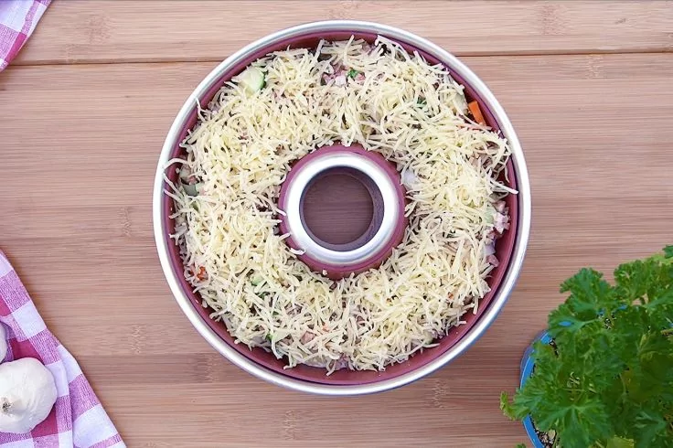 Runde Backform mit Loch in der Mitte, mit geriebenem K&auml;se bedeckt, auf einem Holzbrett; links kariertes Tuch und Knoblauch, rechts ein Topf mit Petersilie